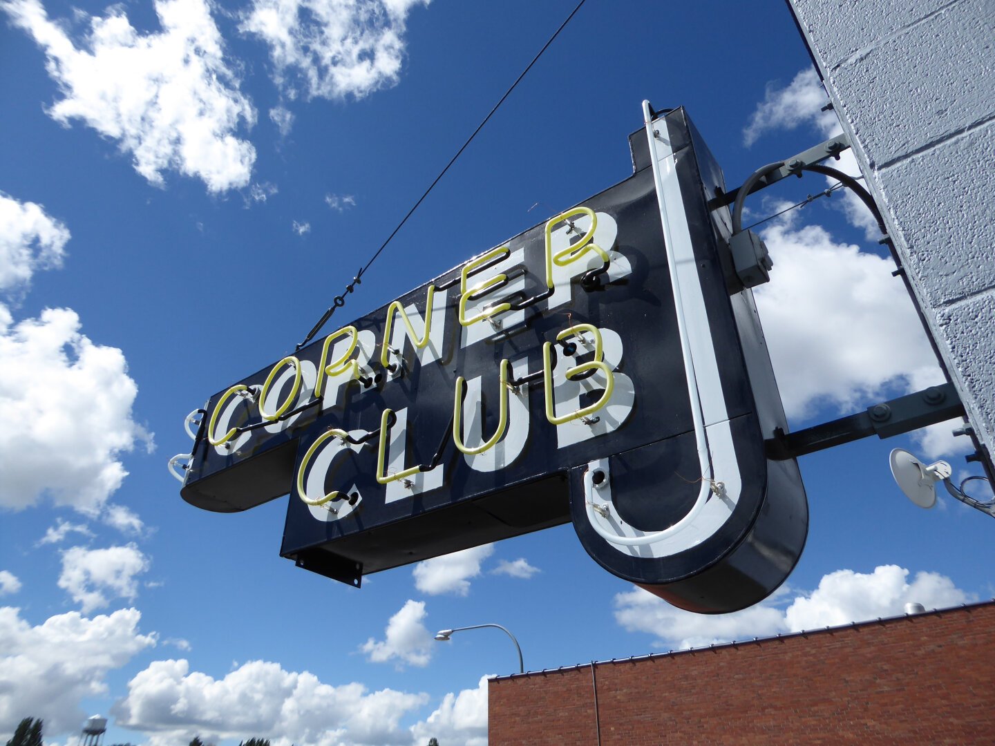 Classic midcentury neon sign against a blue sky with white fluffy clouds. The sign is black, with yellow neon block letters that say Corner Club. There is a backwards J-shaped neon piece to the right.