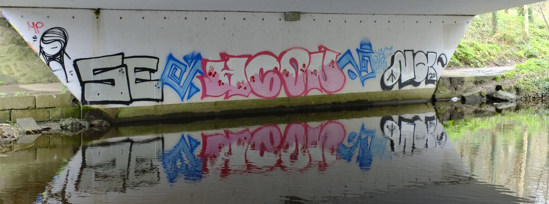 Graffiti on a concrete bridge support reflected in calm water.