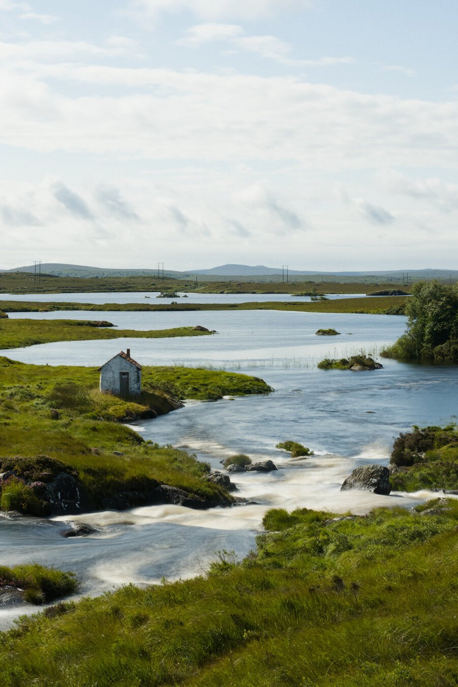 A small, weathered building sits on a grassy bank at the edge of a winding river, under a vast sky with scattered clouds.