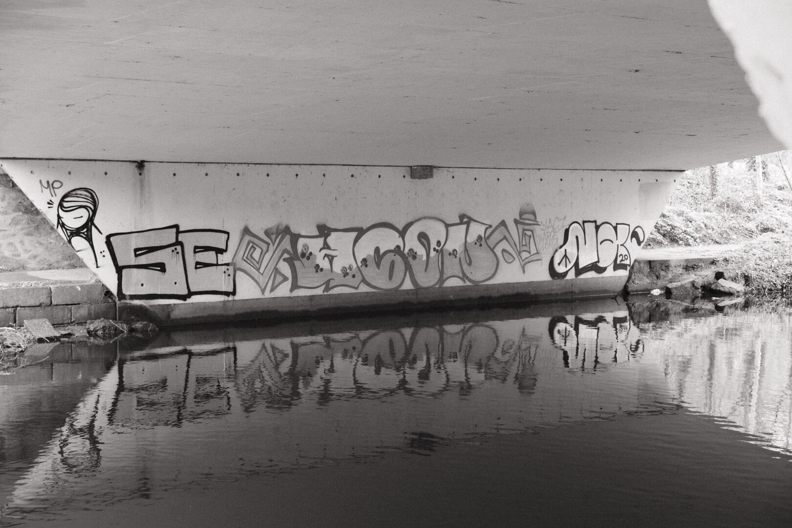 Black and white photograph of graffiti on the underside of a bridge reflected in still water.