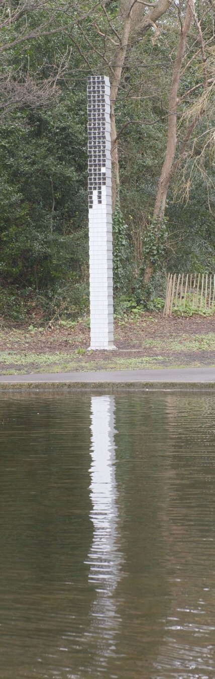 A tall white pillar, some kind of monument or art installation, stands near a pond, reflected in the gently rippling water.