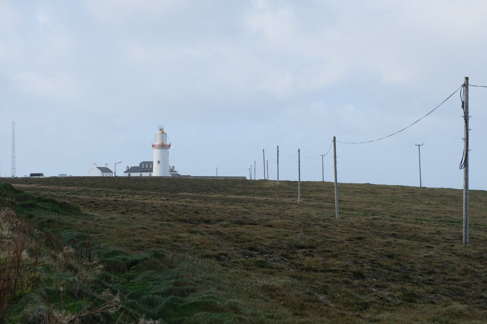 A tall white lighthouse on a hill covered in a green meadow. A series of telephone poles leads in from the right of the frame towards the lighthouse.