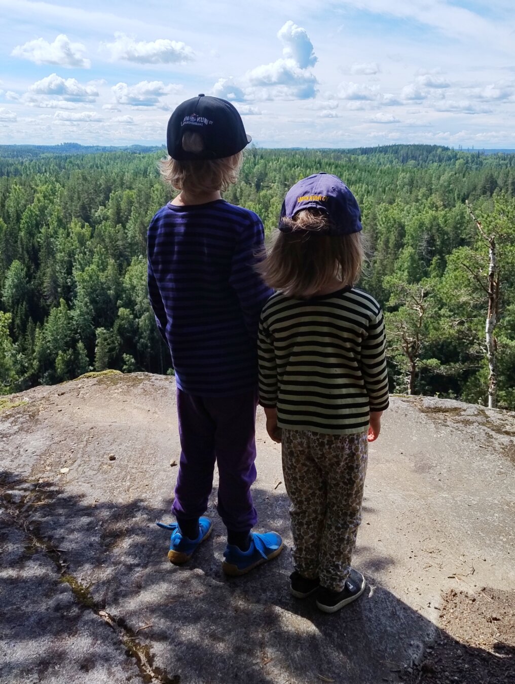 Our kids, 6 and 3, standing on a cliff (a safe distance from its edge), facing a South Karelian forest landscape. Both wear caps that advertise Lapin Kumi, a tyre company their great-grandfather started.
