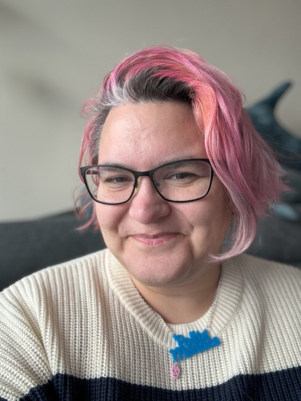 A selfie of Clair, a white person with pink hair, smiling at the camera. They are wearing a blue necklace that has the words “polite menace” on it.