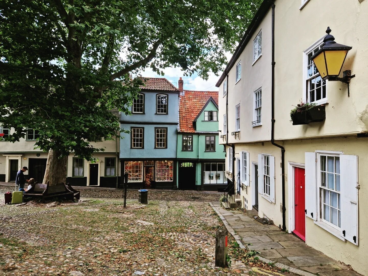 We're on a little square in Norwich. To the right, white buildings with white windows, fairly flat, the one closer to the camera has a red door. There's an old yellow-looking lantern on the nearer building. The floor is cobblestone. In front, behind the little square, two old buildings, very beautiful, one is blue, the other green. To the right, a large tree with benches underneath. Two people are sitting on a bench there, next to luggage.