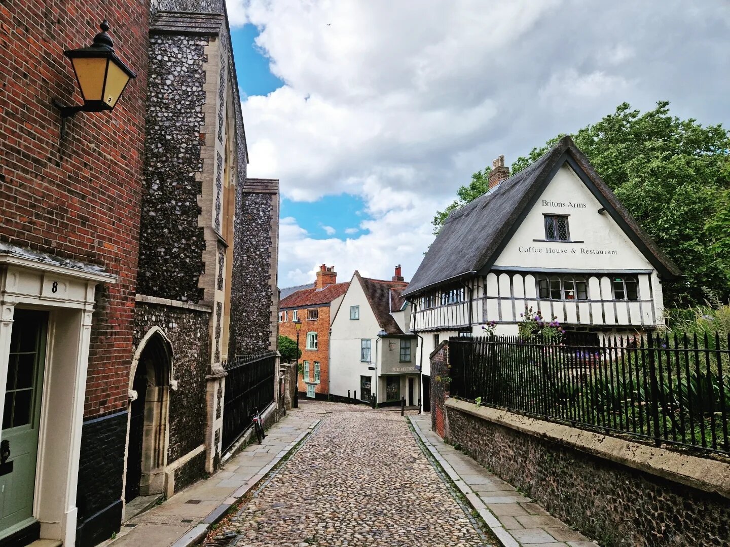 Cobblestone street in Norwich, with an old pub to the right (white, dark brown wooden beams), and old red and grey buildings to the left.