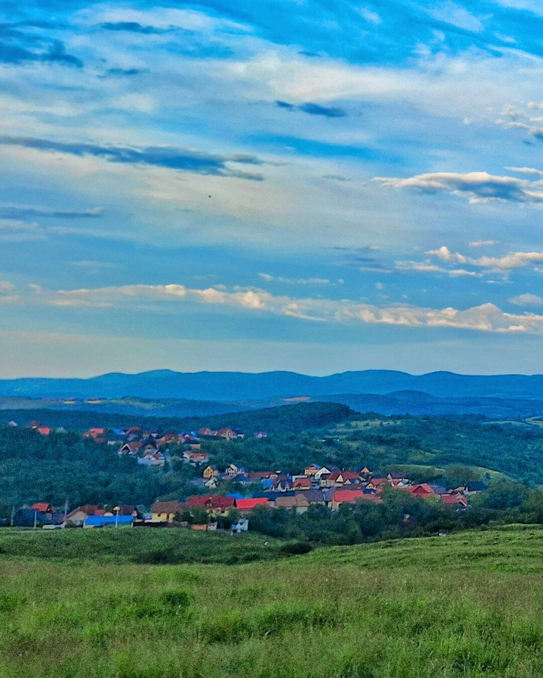 A vibrant aerial view of a Romanian village with red rooftops nestled among green hills, beneath a dramatic blue sky with scattered white clouds and distant mountains.