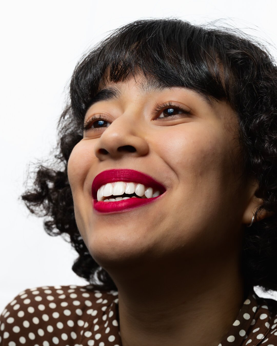 A smiling person with short, curly hair, wearing a brown-and-white polka-dot top. Their bright red lipstick contrasts sharply with the white backdrop. The shot is cropped closely around the face, highlighting the person’s wide smile and cheerful expression.