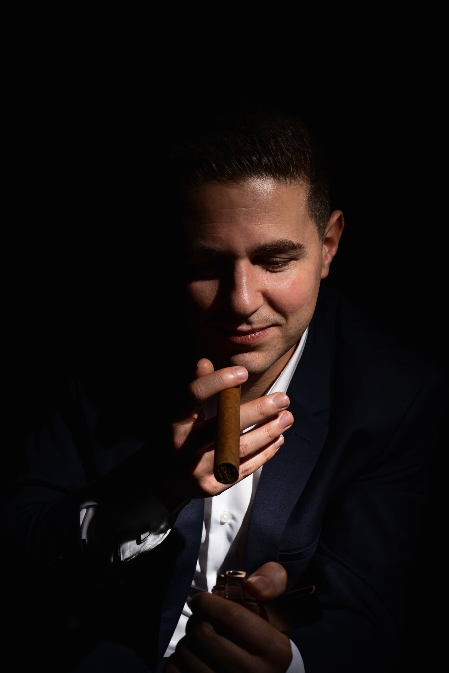 A person in a dark suit jacket and white shirt, holding a cigar near their mouth with their right hand while raising a lighter with their left hand. They are partially lit from the front, leaving the rest of the frame in deep shadow against a black background.