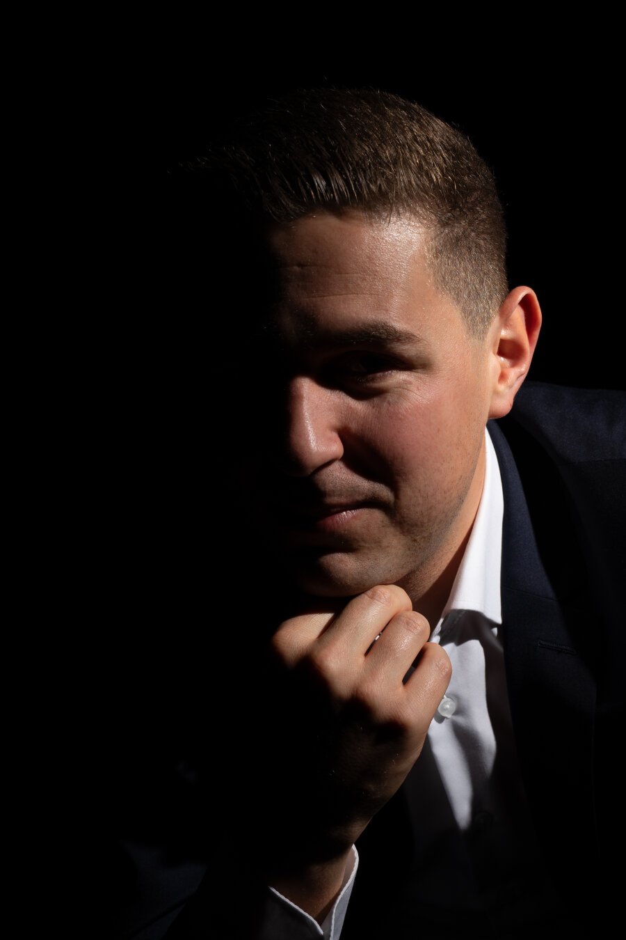 Close-up shot of a person in a dark suit jacket and white shirt, leaning forward with chin resting on hand. The right side of the face is softly illuminated, while the left side remains mostly in shadow against a black background.