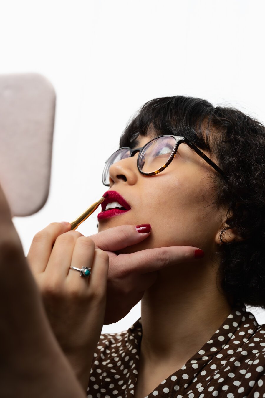 Close-up of a person with short, curly hair and round, dark-framed glasses, tilting their head back slightly. They are applying bright red lipstick with a small brush, supported by another person’s hand beneath their chin. The background is a clean white, drawing attention to the person’s face.