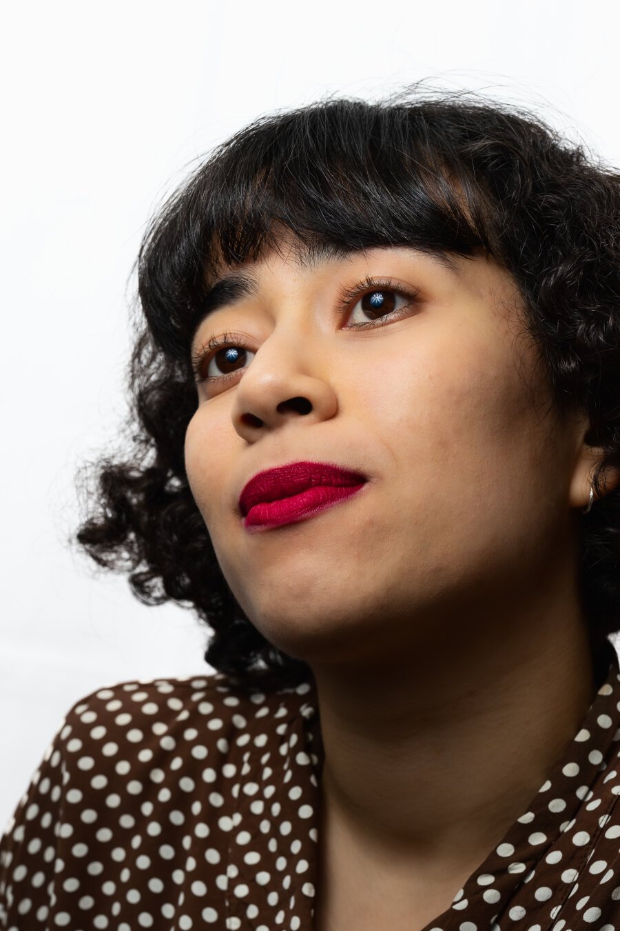 A person with short, curly hair and round, dark-framed glasses, gazing upward. They wear a brown-and-white polka-dot top, and their lips are painted bright red. The plain white background emphasises their facial features and subtle smile.