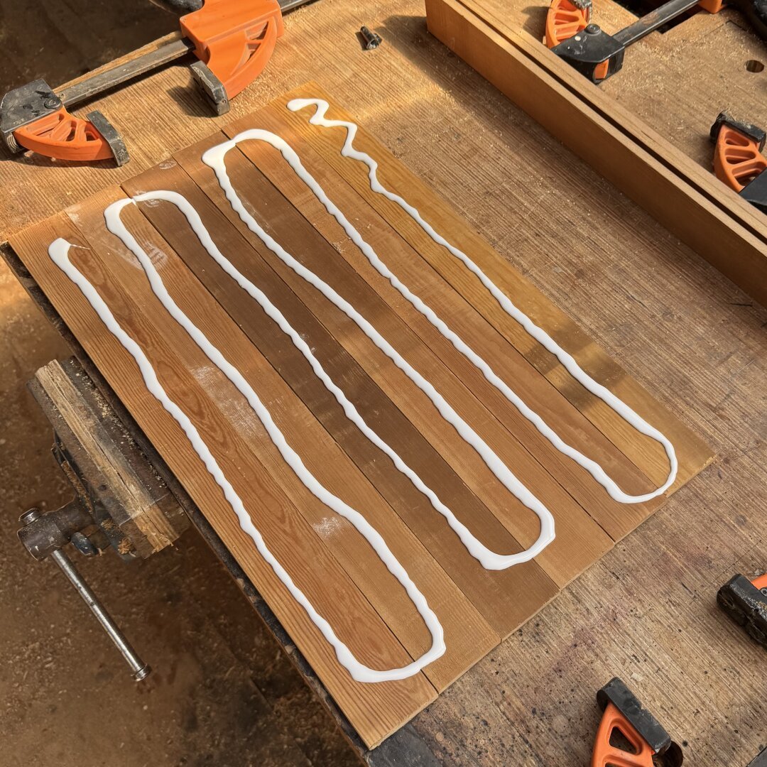 Wooden slats on a workbench with open clamps laying ready next to them. On the slats is a long, thick line of white wood glue, ready to be spread out for gluing up. The light is warm sun with a golden glow.
