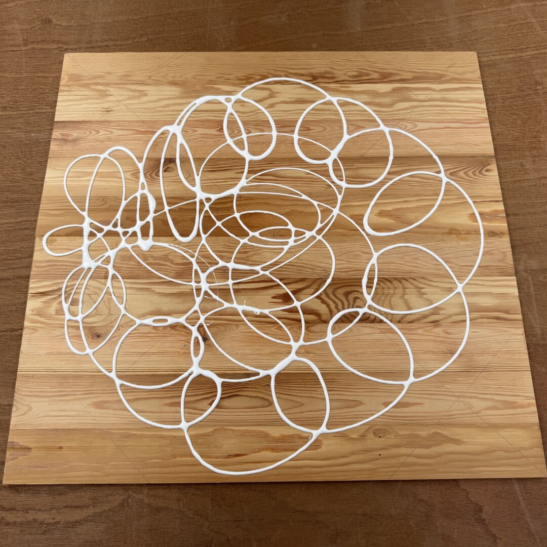 A square board made up of wooden slats on a workbench. On the board is white wood glue in circular lines, ready to be spread out for gluing up. The light is warm sun with a golden glow.