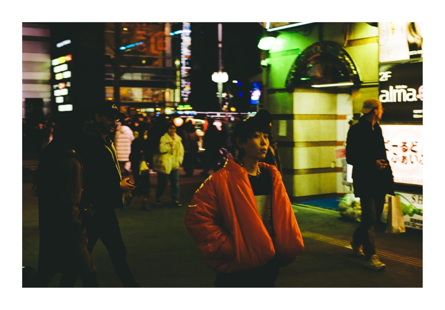 A girl in a red puffy jacket stands on a crowded street at night.