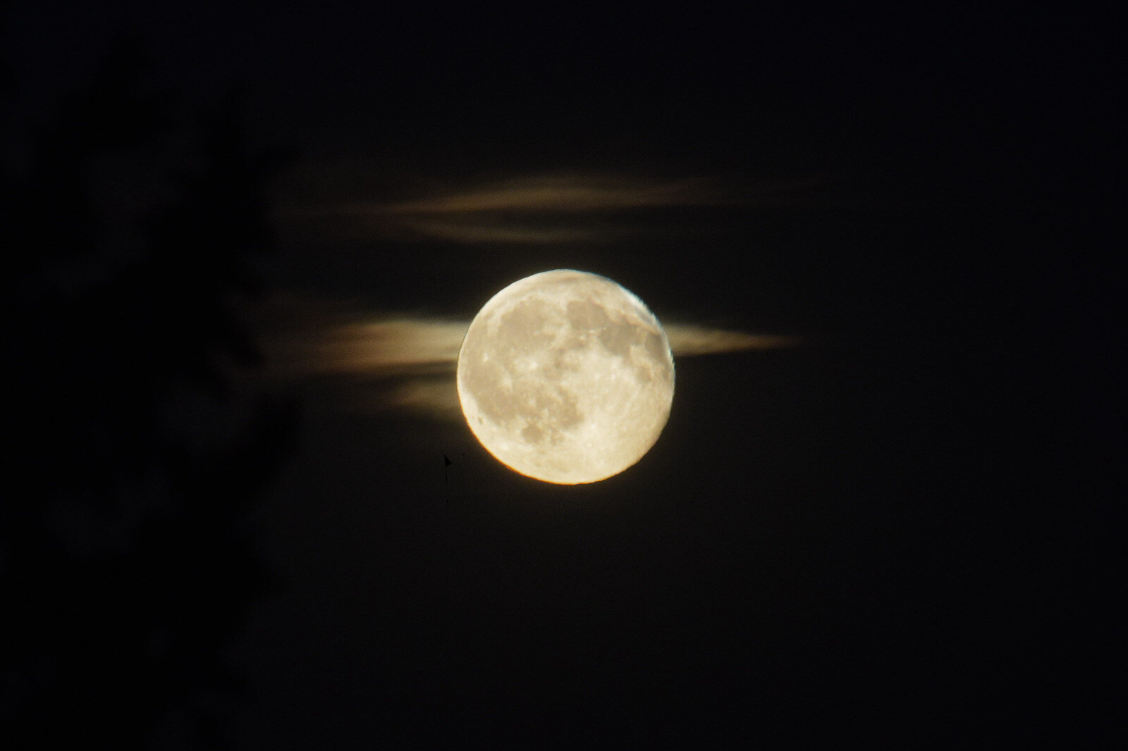 Nearly full moon centered against a dark night sky, with thin wisps of cloud passing in front and faint lunar surface details visible.
