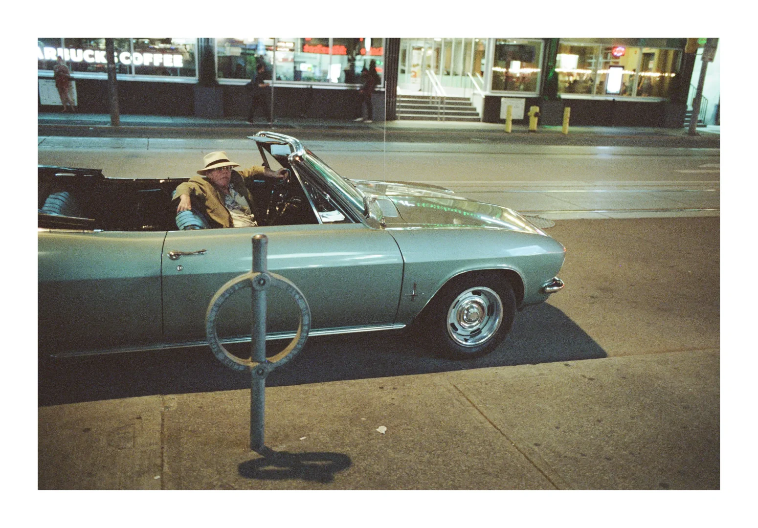 A colour photograph of a man in a convertible. He is wearing a tan sport coat with a Hawaiian print shirt on underneath with a straw fedora and large rim glasses. 

He looks at the viewer with a pose of nonchalance, an arm over the passenger seat back.