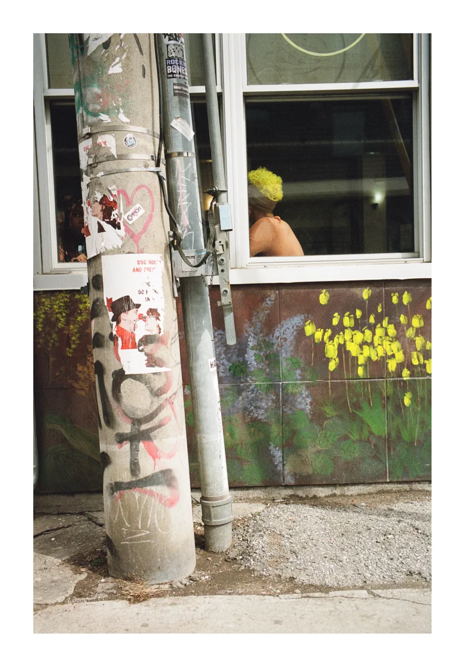 A colour photograph of a woman viewed through a window of a restaurant, her face is obscured by the window frame. She has a shock of dyed electric yellow curly hair. Below the window, the wall of the restaurant has a mural painted with yellow daffodils.