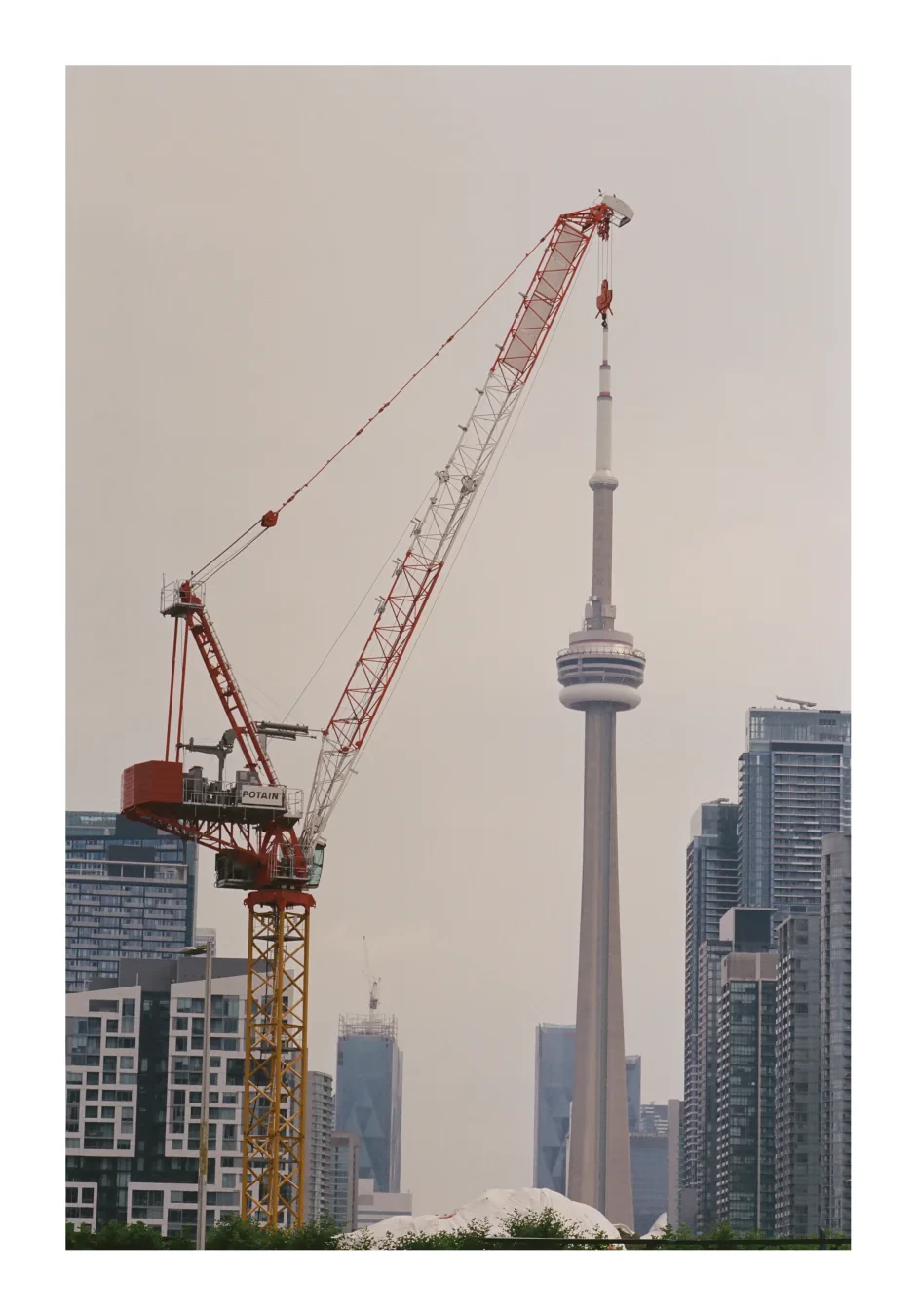 A colour photograph of a construction crane in the foreground with the CN tower behind it. It looks as if the CN tower is being hoisted by the crane.
