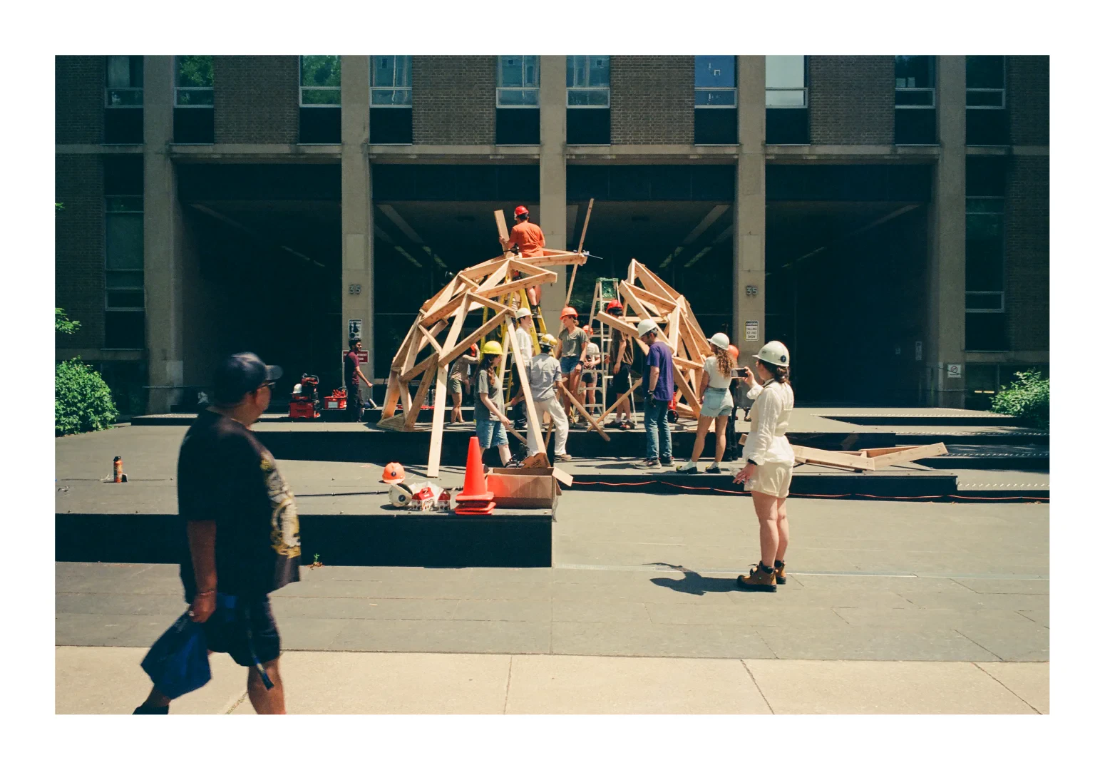 A colour photograph of University of Toronto engineering students assembling a dome structure out of wood. 
The dome is a hive of activity with passers by onlooking.