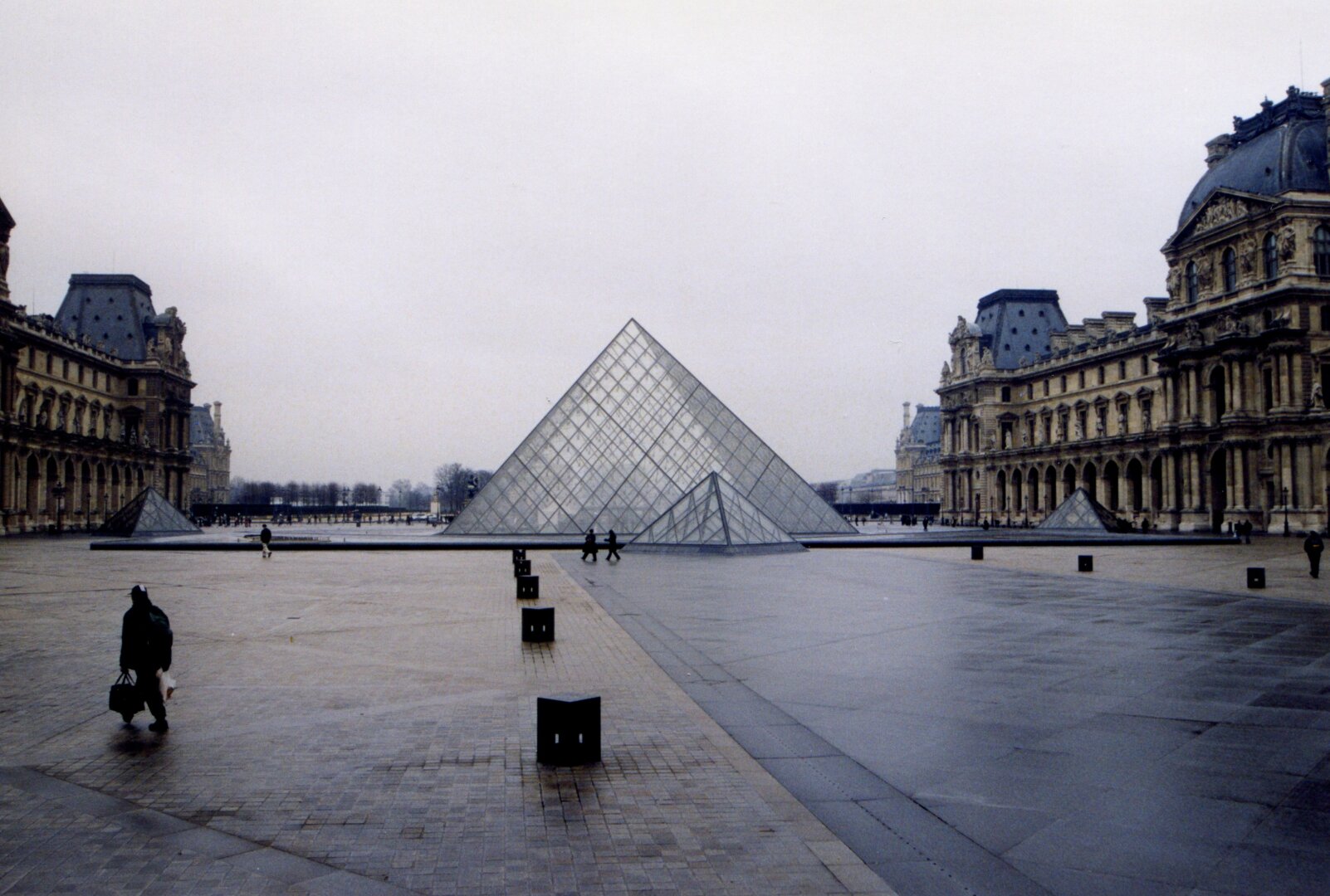 A wide shot of the Cour Napoléon at the Louvre Museum in Paris under an overcast, moody winter sky. In the center of the expansive, wet cobblestone plaza stands the iconic glass and steel Louvre Pyramid, flanked by smaller secondary glass pyramids.

The grand, ornate classical architecture of the Louvre Palace frames both the left and right sides of the courtyard, its detailed stone facades and dark mansard roofs contrasting beautifully with the sleek modern geometry of the glass structure. A few scattered pedestrians dressed in dark winter coats stroll across the quiet, damp square, with soft reflections glinting off the wet pavement. The overall atmosphere is serene, atmospheric, and quiet, capturing a serene moment in the heart of the city.
