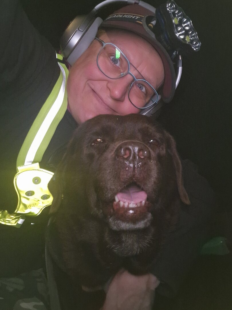 Night time selfie of a person and a chocolate brown Labrador with greying snout. The person is wearing a black jacket, yellow safety harness, red cap, silver headphones and blue-rimmed glasses. On their cap rests a headstrap torch in off position. Their free hand is around the dog's shoulders. Both are facing the camera. The dog's mouth is slightly open, showing off some blep.