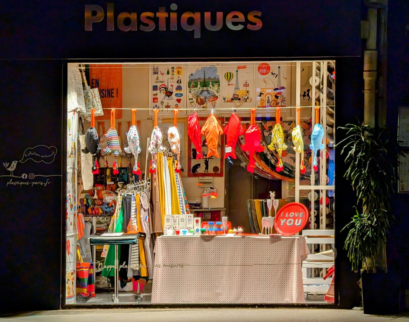 A storefront at night. Various colored items are visible, amongst which a collection of fake chickens.