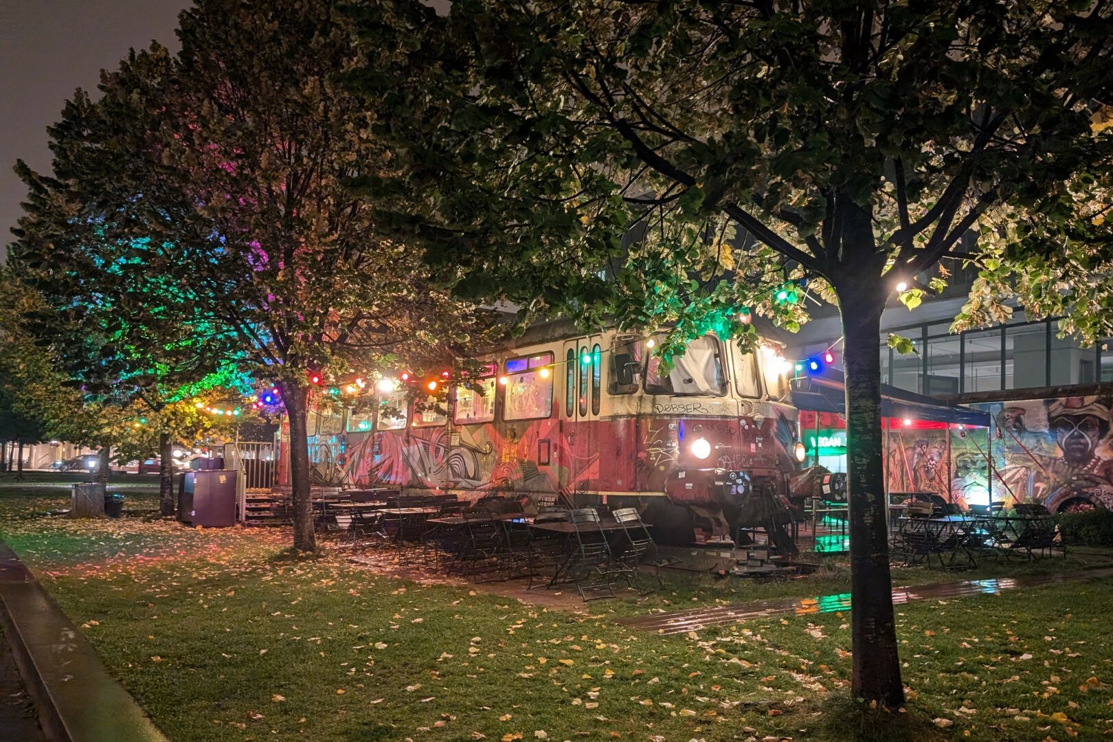 An old red métro car has been turned into a bar: on the grass, surrounded with tables and trees, it stores drinks and foods. The photography was taken at night, many colored lights hanged in the trees give an eery feeling to the place.