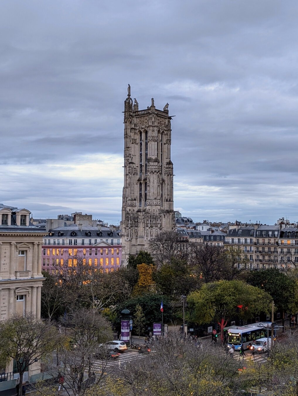 View of the tower Saint Jacques in Paris from the top floor terasse of a theatre next by. Below the tour one can see the trees of the garden surrounding the tower. Behind, the façades of the buildings are lit in pastel colors. The sky is grey from the falling night behind the clouds.