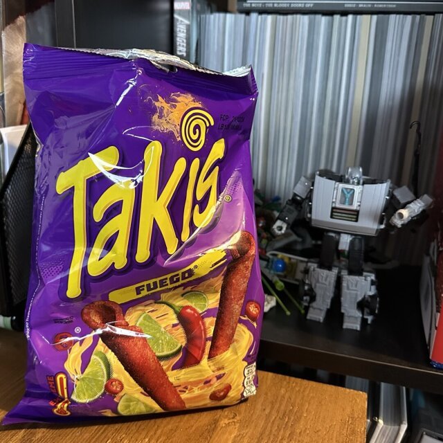 A bag of Takis Fuego on a desk. Behind it is a bookshelf with a Delorean transformer.