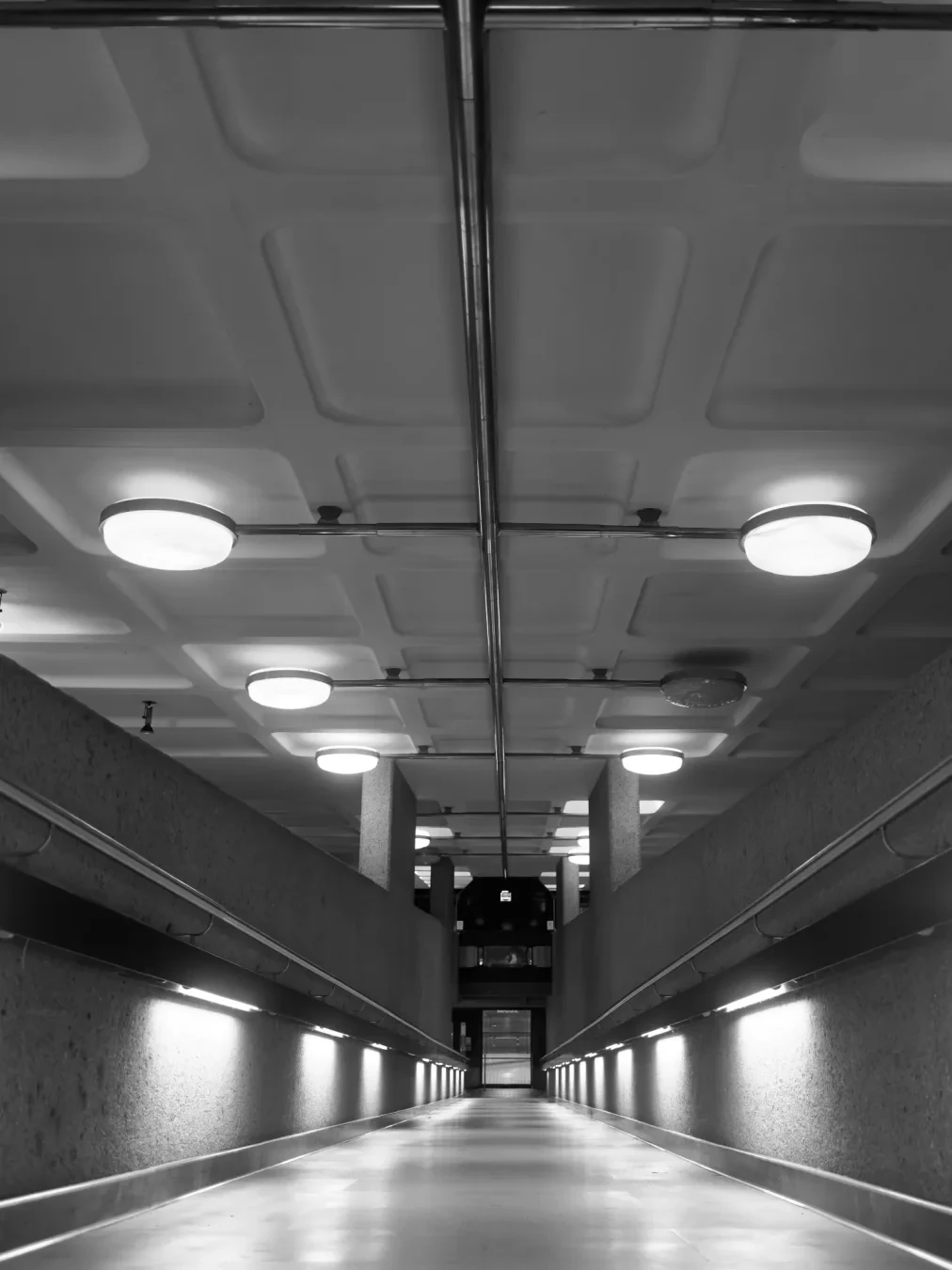 Smooth ramp going down inside of the building with lights and handrails on both side. Ceiling is just painter concrete with geometric shapes