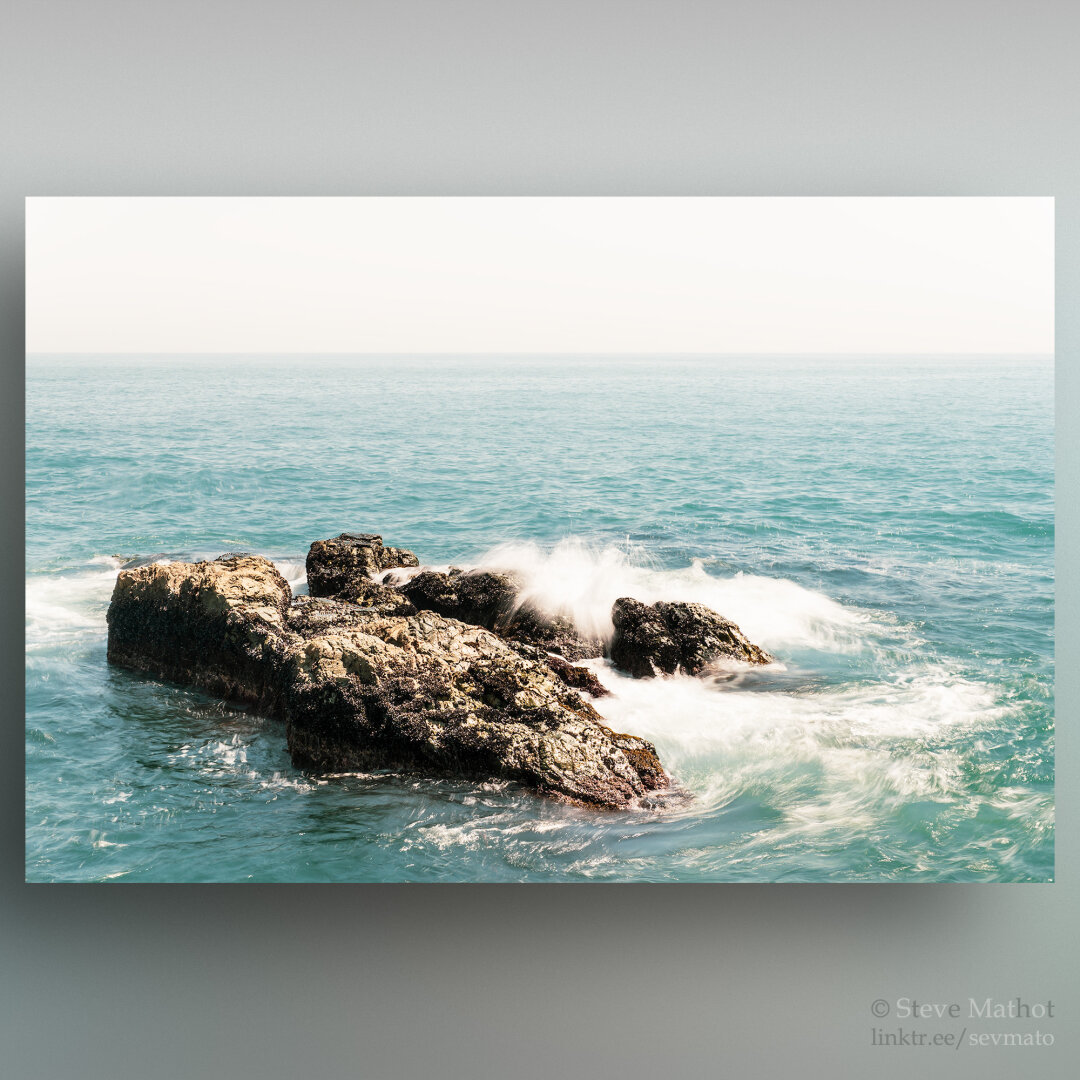 This photo showcases the vibrant turquoise waters of the ocean as waves crash against rugged rocky outcrops. The partially submerged rocks add a dramatic element to the scene, with the spray from the waves creating a misty, dynamic effect. The horizon merges seamlessly with the hazy sky, enhancing the sense of vastness and natural beauty.