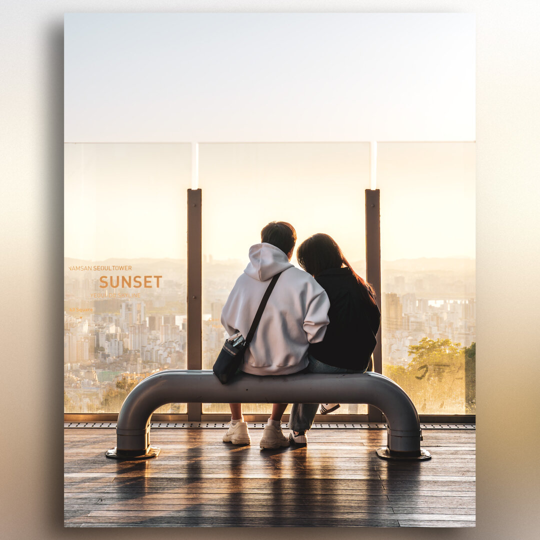 A couple sit closely together on a curved metal bench at sunset, overlooking the Seoul cityscape from Namsan Seoul Tower. The man wears a white hoodie with a black crossbody bag, the girl a dark jacket. In front of them, a glass barrier displays text about the skyline, and the hazy, golden light bathes the buildings below in a peaceful glow.