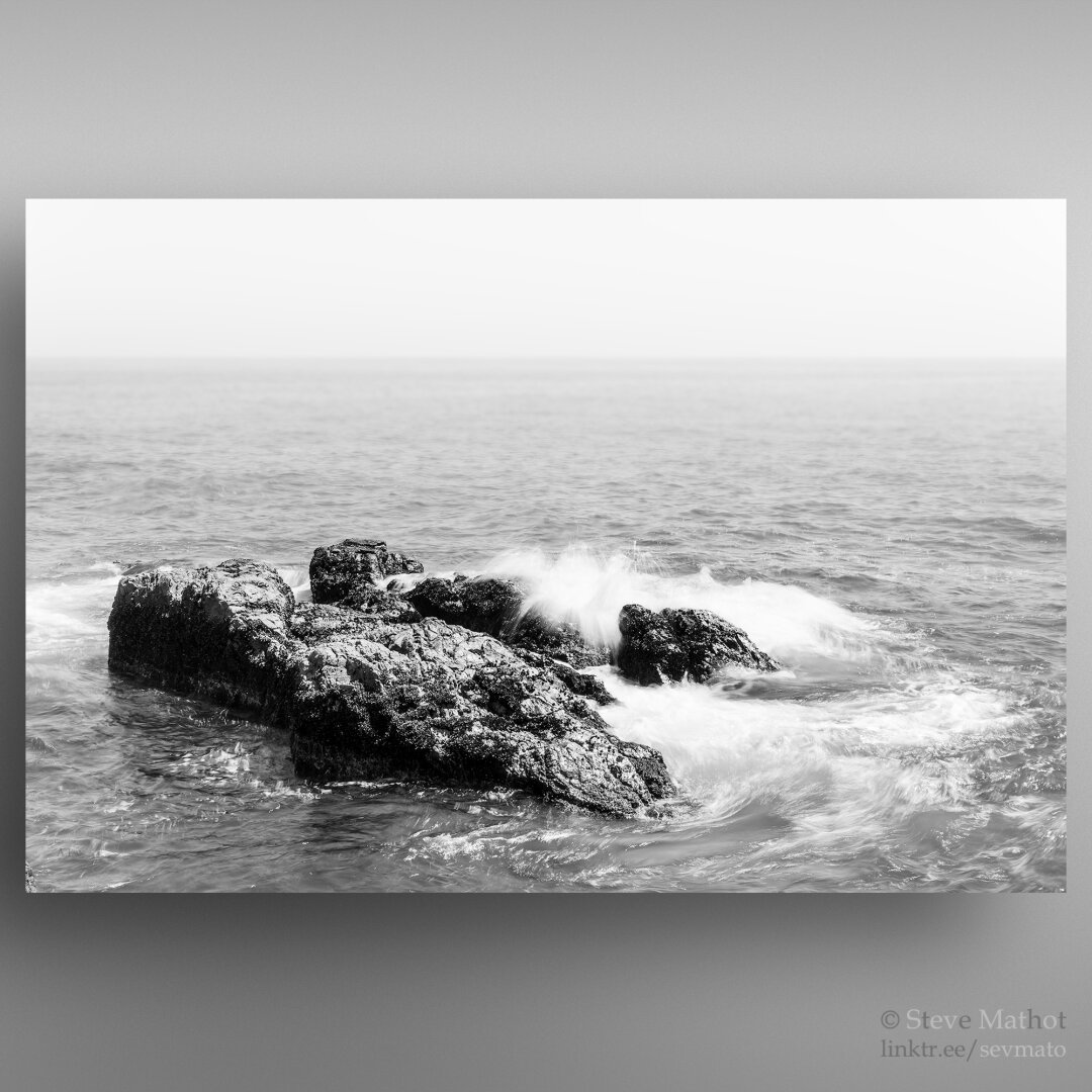 This photo (Black and White) showcases the vibrant turquoise waters of the ocean as waves crash against rugged rocky outcrops. The partially submerged rocks add a dramatic element to the scene, with the spray from the waves creating a misty, dynamic effect. The horizon merges seamlessly with the hazy sky, enhancing the sense of vastness and natural beauty.