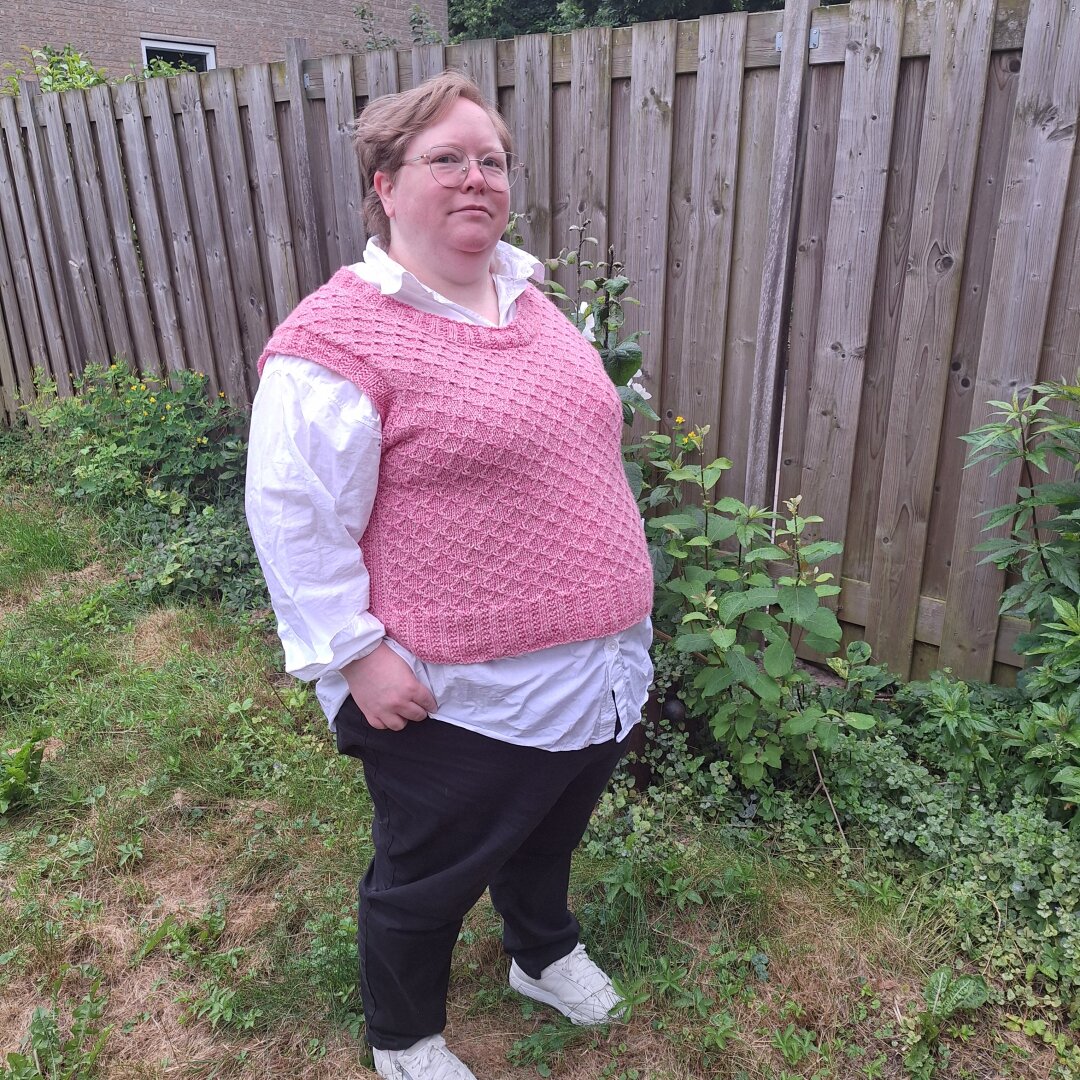 Person standing in a garden, wearing white shoes, black pants, a white button down shirt and a pink textured vest