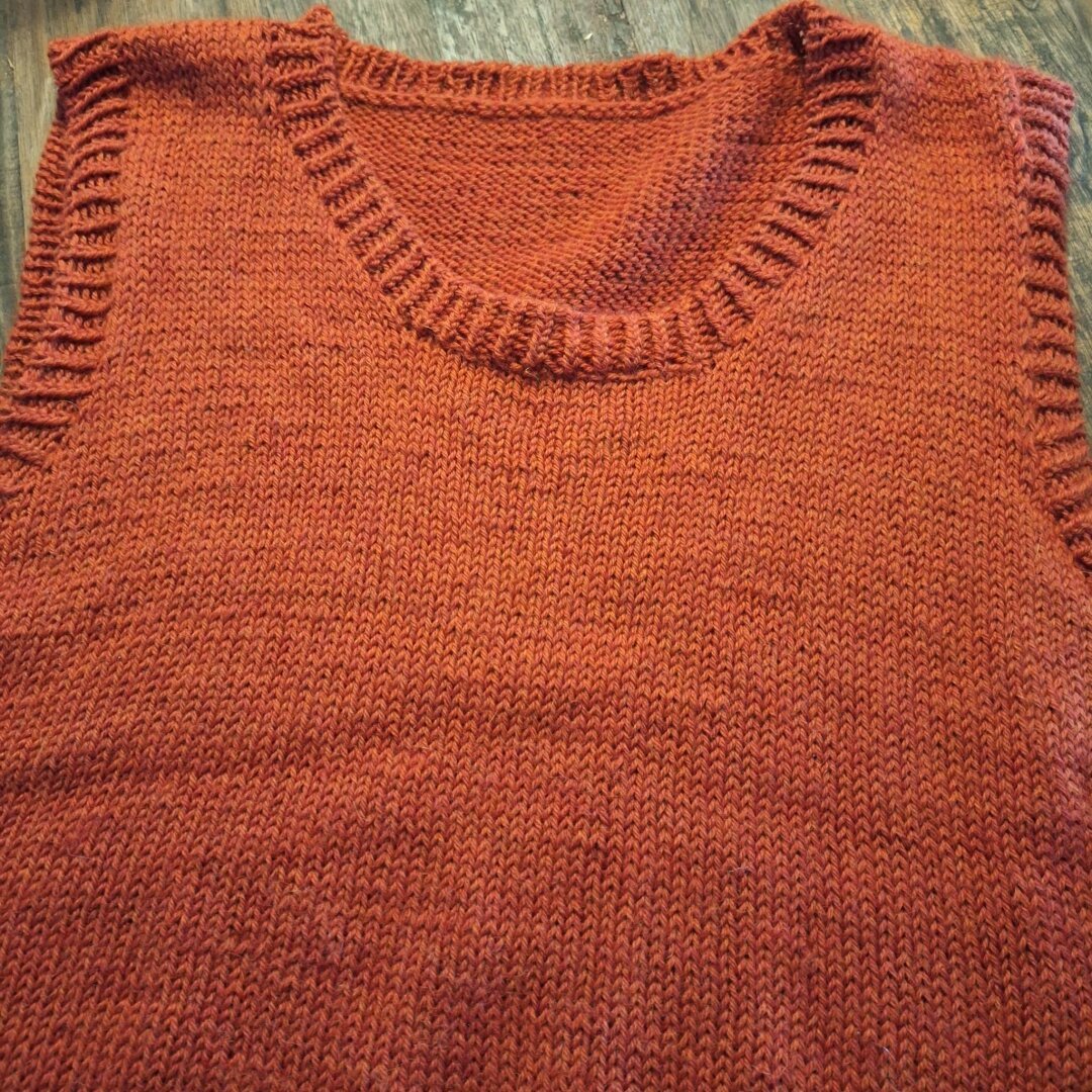 A red/orange knitted vest in a slightly fluffy yarn on a dark wooden table.