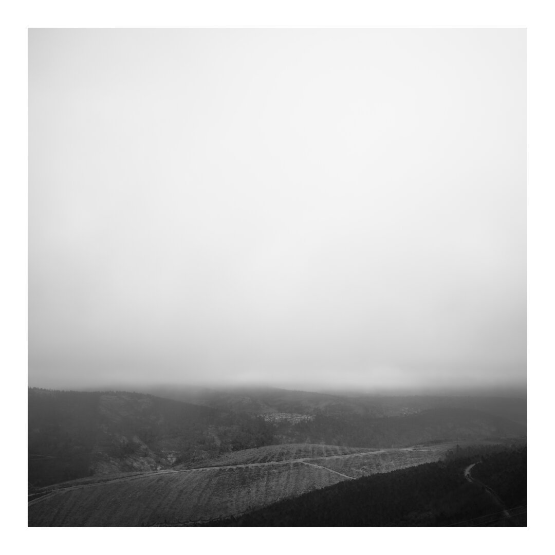 A monochrome landscape of hills fades into a foggy abyss. The objects in the background are obscured, except for the faint outline of structures clinging to the hillsides. The only path is a solitary road winding its way down the mountain.