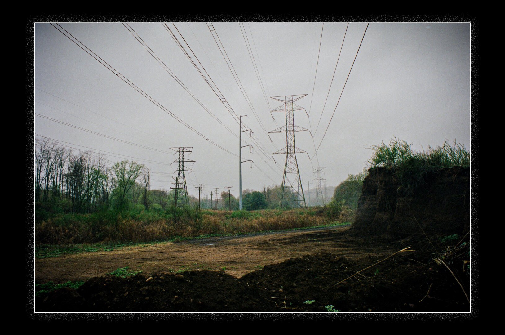 A landscape featuring multiple high-voltage power lines and transmission towers stretching across a cloudy sky. The ground below is a mix of dirt paths, vegetation, and shrubs, with a wooded area in the background. The atmosphere is overcast and damp, suggesting a misty or rainy day.