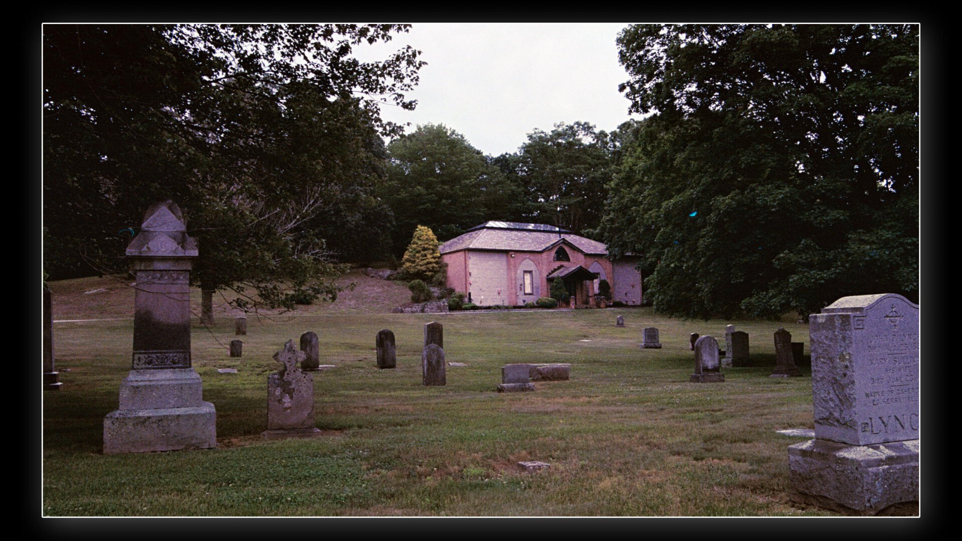 A cemetery with weathered gravestones scattered across a grassy field, surrounded by trees. In the background, there is a small pinkish-red chapel-like building with a pitched roof and arched windows. The overall atmosphere is serene and slightly overcast