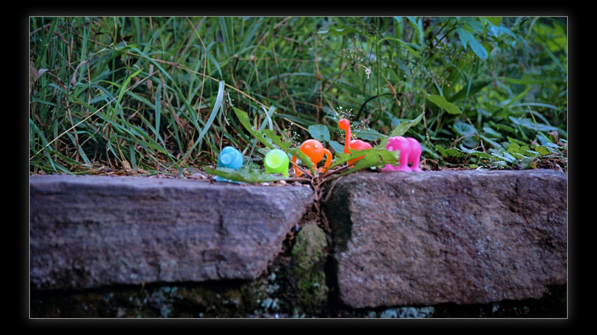 A close-up photograph of colorful plastic figurines resembling snails and animals placed on a stone surface surrounded by lush green grass and plants in the background.