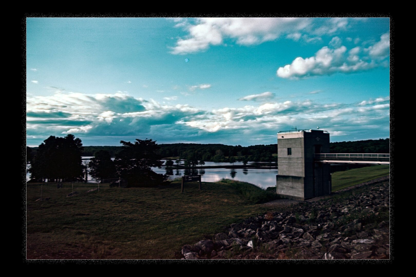 A scenic view of a dam with a concrete structure on the right, overlooking a calm body of water surrounded by lush green trees. The sky above is bright blue with scattered clouds, creating a peaceful and natural atmosphere.