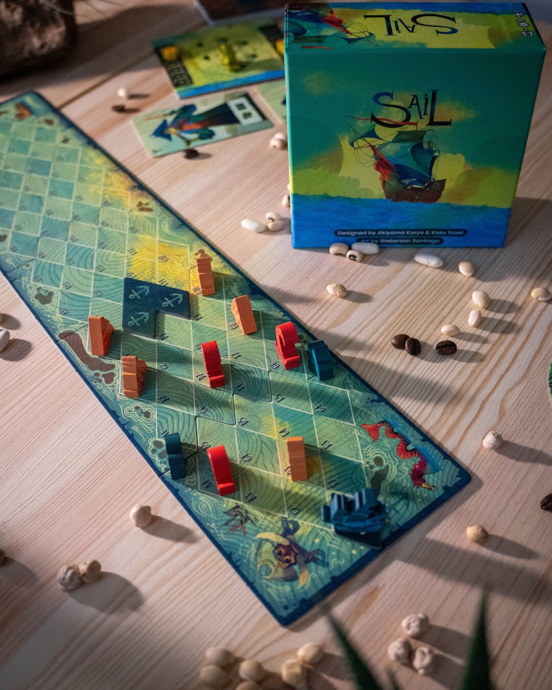 The board game Sail, displayed with the game board and pieces from the box, on a wooden table with an assortment of beans surrounding it.