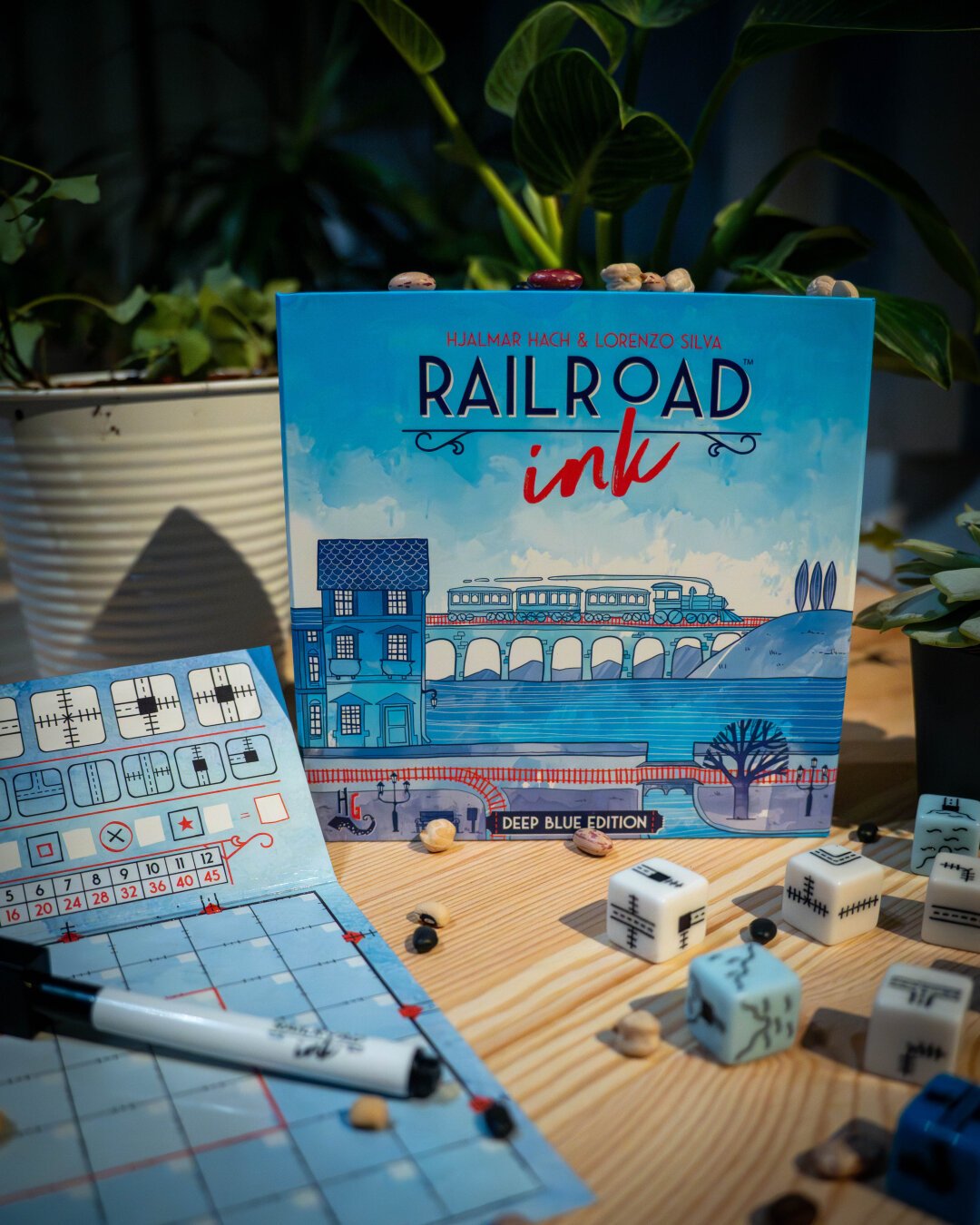 The board game Railroad Ink displayed on a wooden table surrounded by plants and various beans. The box art features a blue painterly scene of a train passing through an environment and the games name. The games whiteboard grid and dice are visible in the photo.