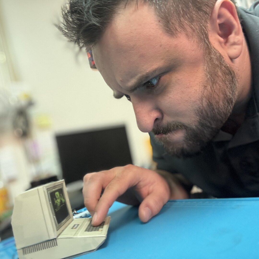 A man with a faux hawk and short beard pretends to be playing on a tiny paper craft Apple 2 computer. The screen on the computer is a screenshot from the original Oregon Trail game in all its monochrome green-on-black glory, showing a covered wagon with oxen, a man, a woman, and two children, a boy and a girl. A text prompt is asking, 