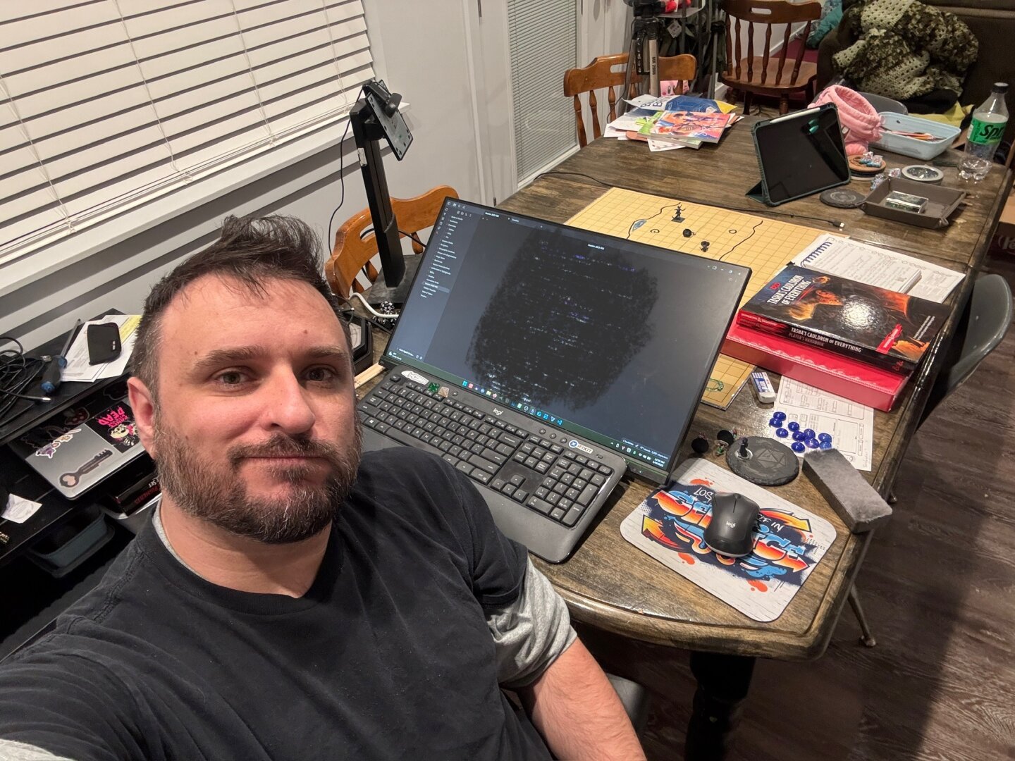 Selfie of a man in front of a table. There's a keyboard, mouse, and monitor; a couple of webcams; and assorted Dungeons & Dragons paraphernalia, such as books, dice, and miniatures on a gridded battle mat.