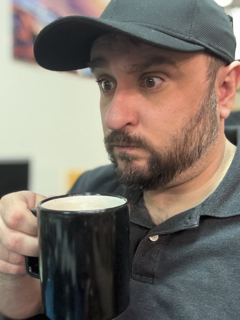 A man wearing a dark gray polo shirt and black baseball cap holds a cup of coffee close to his face while staring out of frame to the left, his face conveying shock and disbelief at what he is seeing.