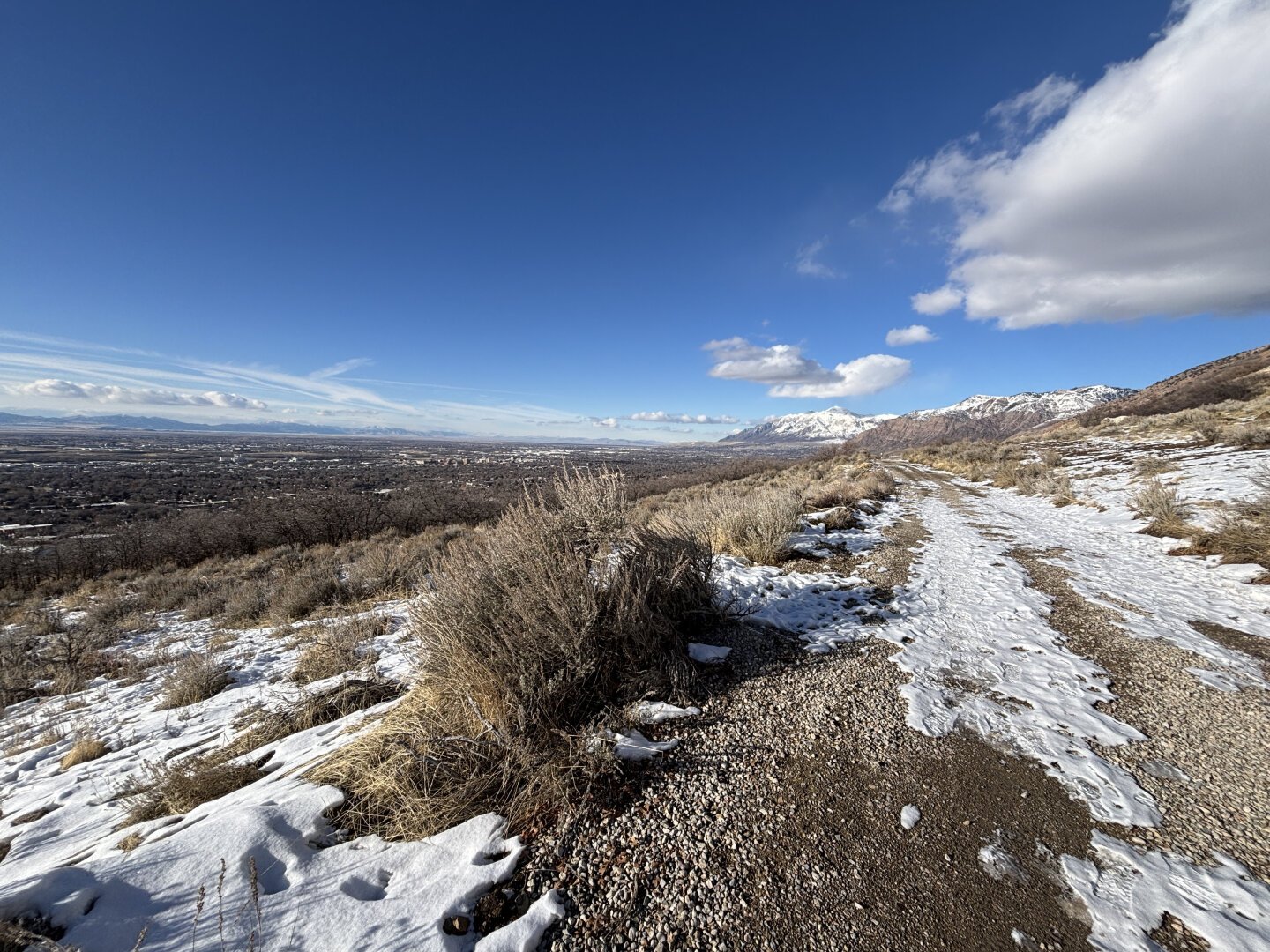 A trail above Ogden, Utah, looking north. The trail is actually an access road so is wide enough and has tracks for vehicles to make their way along the mountain side. It is covered with some residual snow and ice.