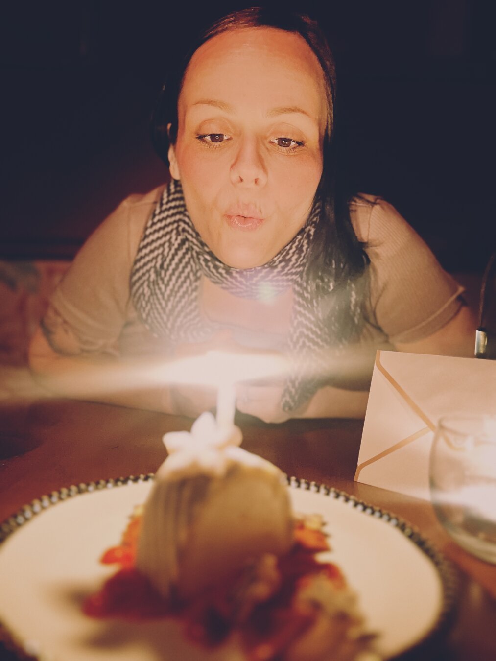 A woman is blowing out the candle on a dessert while wearing a scarf. The setting is dimly lit, highlighting the candle's flame and the dessert on a plate, which has some decorative elements beside it. An envelope is partially visible in the background.