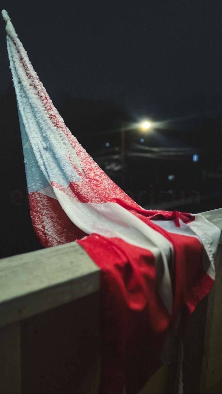 A weathered Puerto Rican is draped over a railing, partially covered in snow. The scene is set at night, with dim lighting in the background from nearby street lamps.