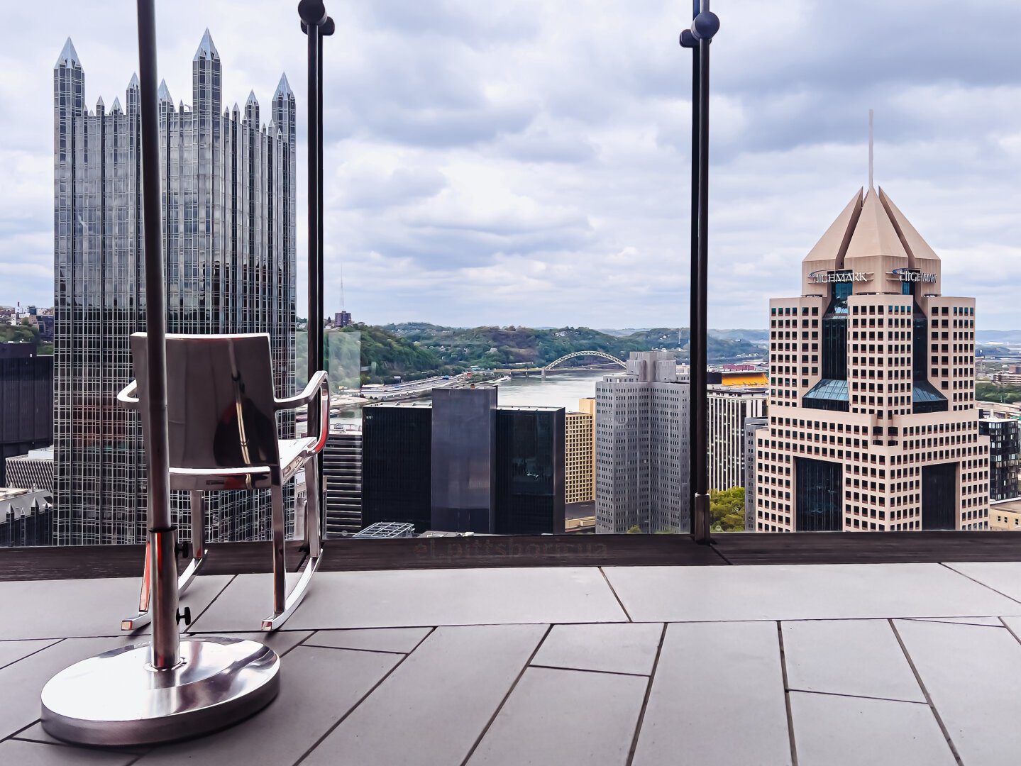 View of city skyline with modern skyscrapers, one prominently featuring a slanted roof, as seen through railings from an elevated terrace with a single chair in the foreground.